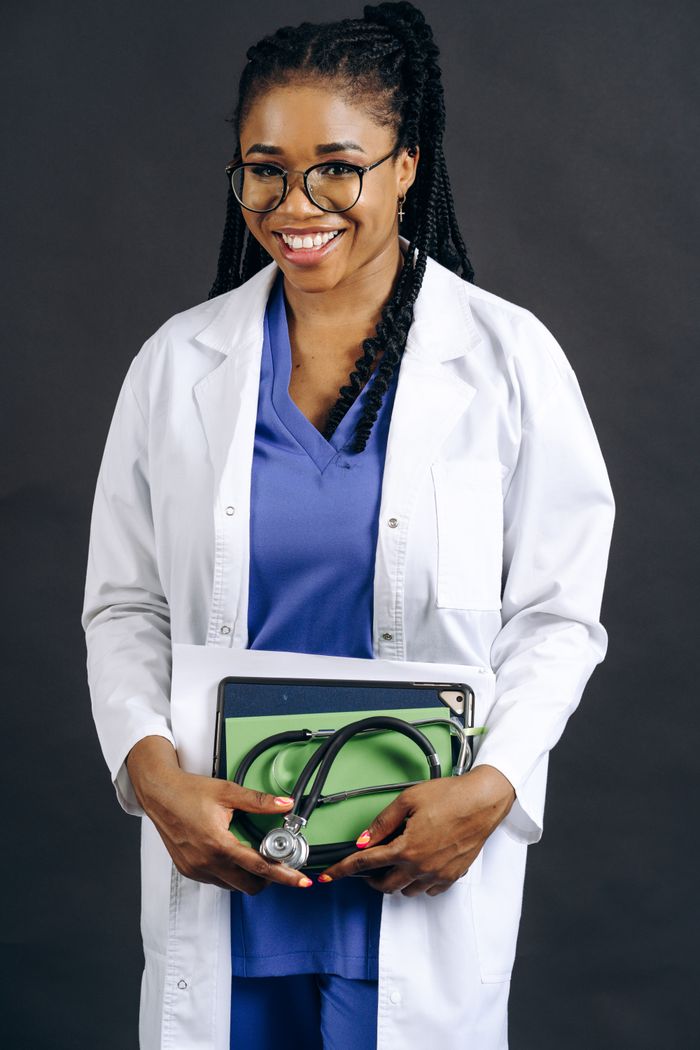 services-02 Portrait of a smiling female doctor in a lab coat holding a stethoscope, against a gray background.