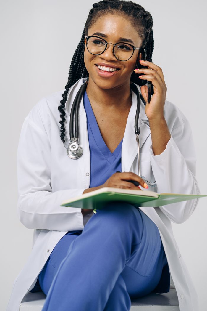 about-bg Black female doctor in medical attire, smiling and talking on a smartphone, holding a notebook.