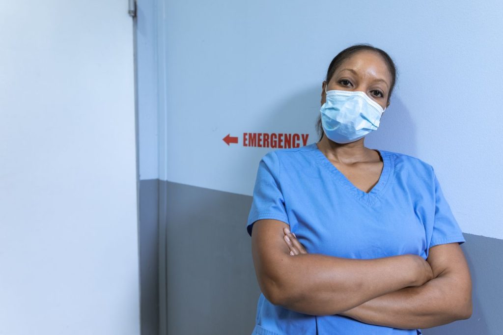 pexels photo 6129199 Nurse in blue scrubs and face mask in hospital corridor with emergency sign.