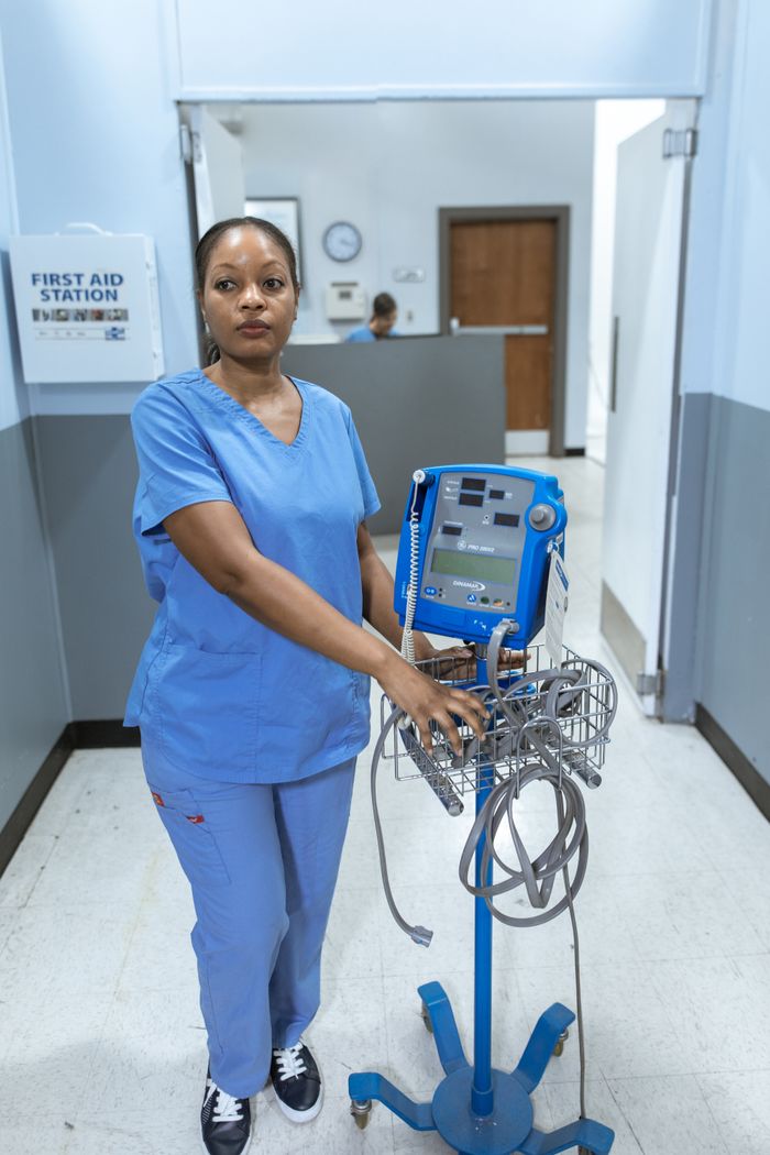 who-we-are A nurse in blue scrubs stands confidently with medical equipment in a hospital corridor.