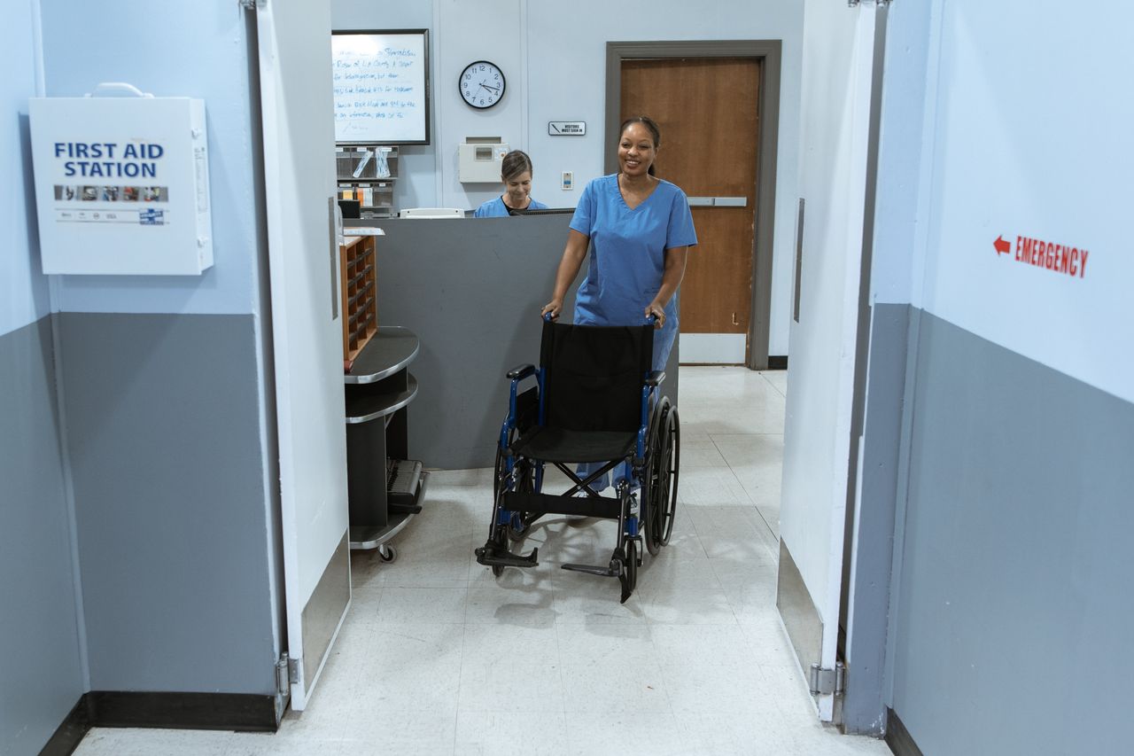 our-story A nurse in scrubs pushes an empty wheelchair through a hospital hallway.